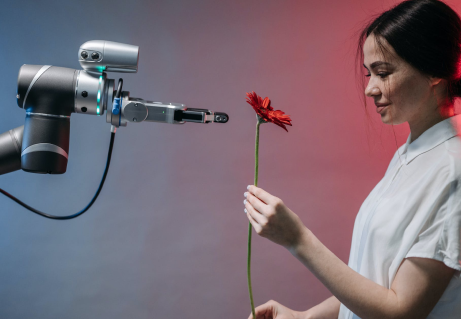 A woman gently receiving a red flower from a robotic arm, illustrating human-robot interaction.