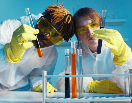 Two scientists in lab coats and safety goggles closely inspecting colorful liquids in test tubes