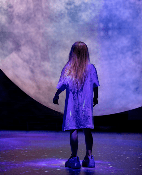 A child silhouetted against a large, luminescent projection of the moon in a dark room.