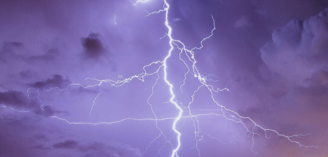 A dramatic lightning strike illuminating the night sky with branching electric bolts, set against a backdrop of stormy purple clouds.